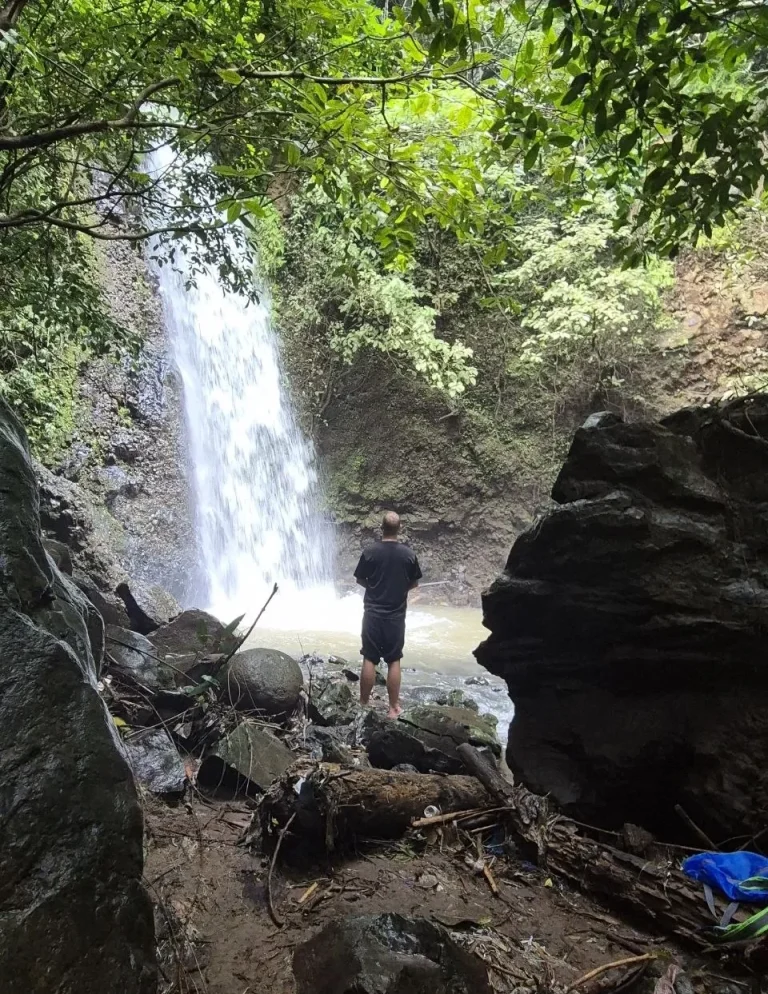 Mann steht vor einem Wasserfall in einer bewaldeten Umgebung.
