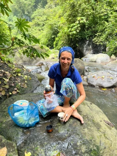Eine Frau in blauer Kleidung kniet an einem Wasserfall und hält Flaschen in den Händen.