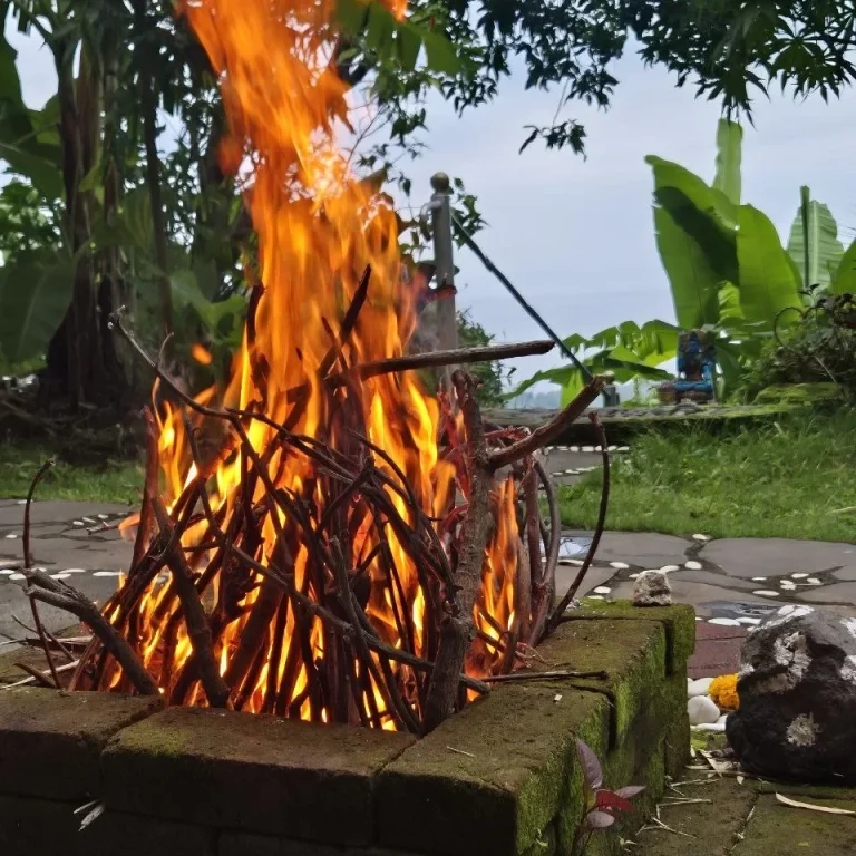 Lagerfeuer mit flammenden Holzscheiten, im Hintergrund Steine und eine Person im Sitzen.