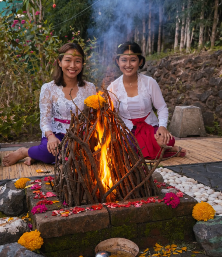 Zwei Frauen in traditioneller Kleidung sitzen neben einem Feueraltar, umgeben von Blumen.