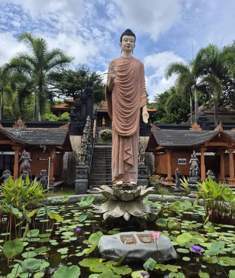 Statue eines stehenden Buddha vor einem Teich mit Lotusblumen und tropischer Vegetation.